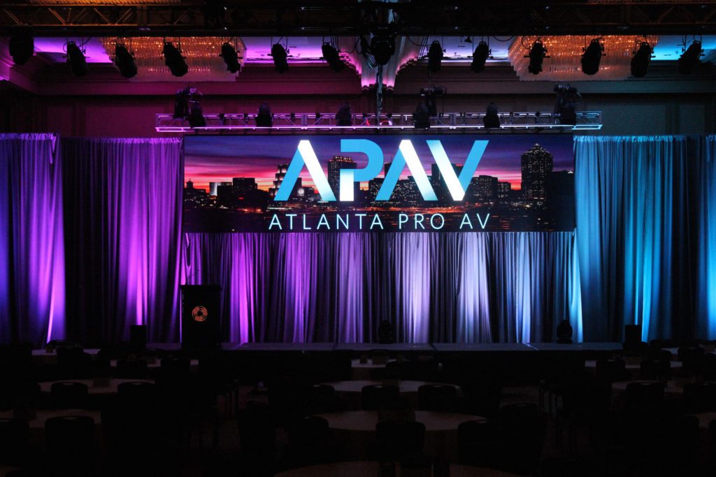 Request a Quote: APAV logo on a cityscape screen in front of curtains under purple and blue lights