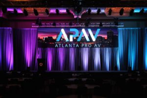 APAV logo on a cityscape screen in front of curtains under purple and blue lights