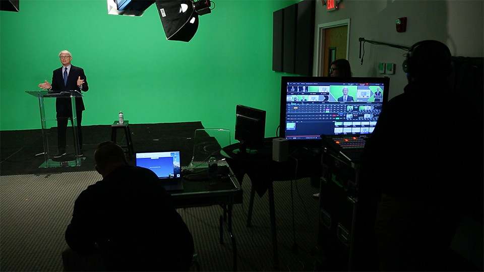 A person presents in front of a green screen while the tech crew watches on monitors.